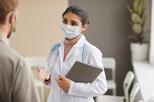 Young female doctor in protective mask holding medical cards in her hands and talking to patient standing at the corridor