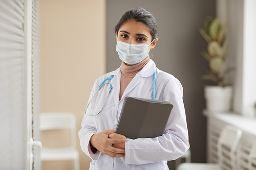 Portrait of female doctor in protective mask and in white coat holding medical cards in her hands looking at camera at hospital