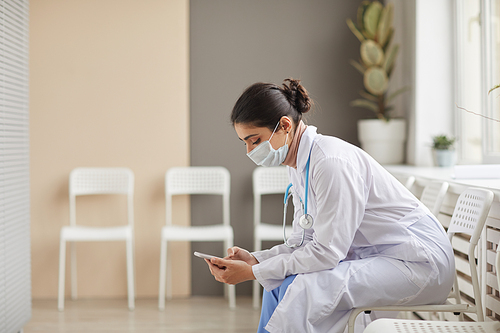 Female doctor in white coat and in mask typing a message on her mobile phone sitting at the corridor