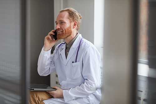 Confident doctor in white coat has a conversation on mobile phone while sitting at the corridor