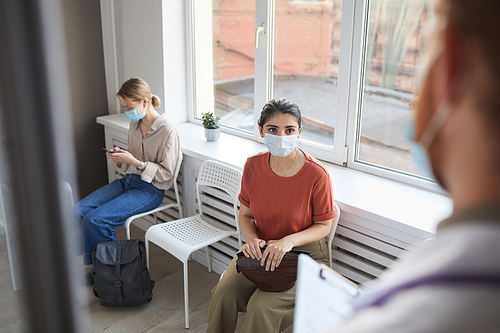 Young woman in protective mask sitting in queue at hospital while doctor meeting her