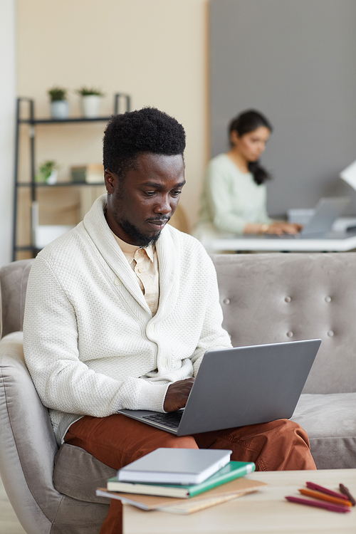 African man sitting on sofa in front of the table with books and typing on laptop he learning online at home