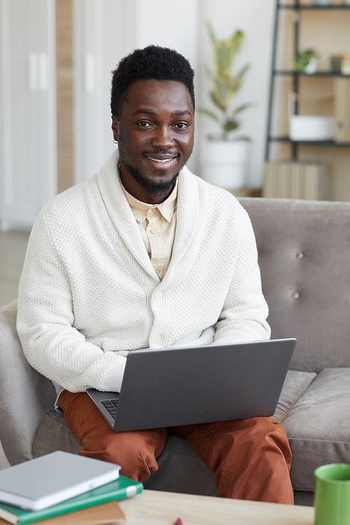 Portrait of African young man smiling at camera while sitting on sofa and working on laptop