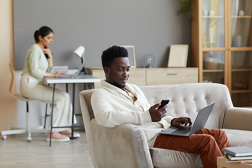 African man sitting on sofa with laptop and using mobile phone with woman working in the background at home