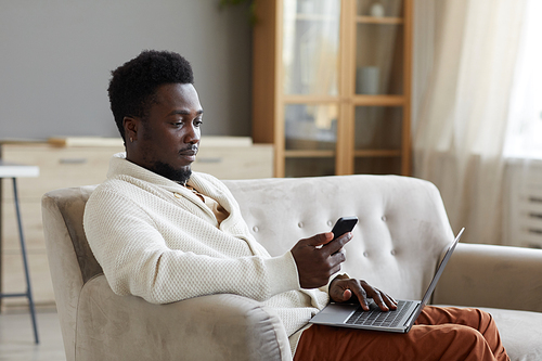 African man reading a message on mobile phone while working online on laptop on the sofa in the room