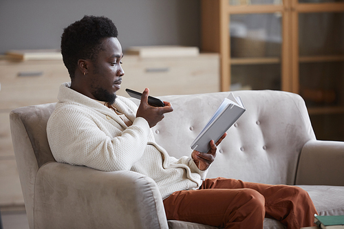 African man sitting on sofa reading a book and recording an audio message on his mobile phone he sitting in the room