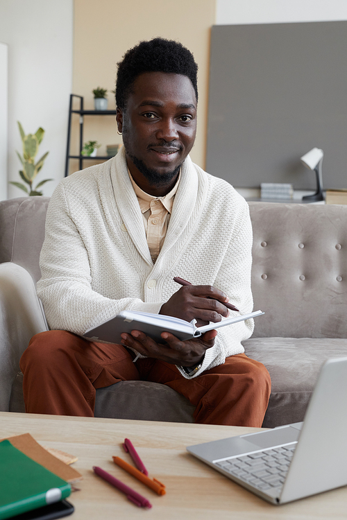 Portrait of African young man smiling at camera while sitting on sofa with books he studying at home