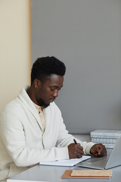 African man sitting at his workplace with laptop and writing business plan in note pad