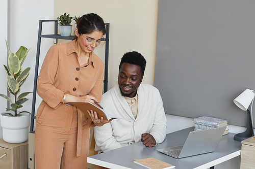 Business partners cooperating together at office African businessman sitting at the table with laptop while businesswoman showing him the document