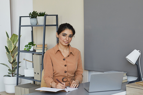 Portrait of young businesswoman sitting at her workplace with laptop and making notes in her note pad she working at office