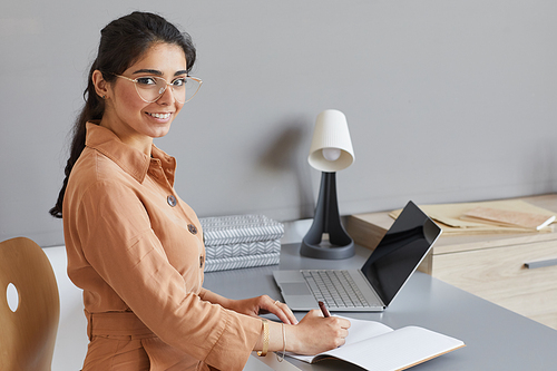 Portrait of young businesswoman smiling at camera while sitting at the table at office and writing plans in note pad