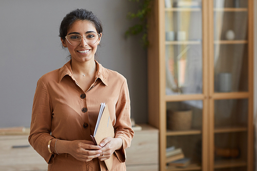 Portrait of young businesswoman in eyeglasses and in blouse holding folders and smiling at camera at office