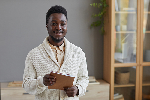 Portrait of young African businessman in white sweater holding folder and smiling at camera standing at office