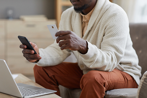 Close-up of African businessman using mobile phone and credit card for online payment sitting on sofa in front of laptop