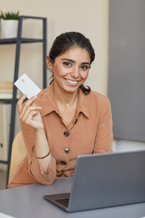 Portrait of young woman smiling at camera while sitting at the table with laptop and buying purchases online with credit card