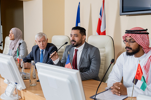 Confident mature delegate or politician making speech while sitting by table among his foreign colleagues at summit