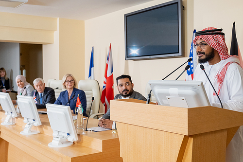 Young confident Arabian speaker in traditional clothes standing by tribune and speaking in microphone at political conference