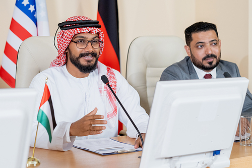 Young confident Arabian delegate speaking in microphone at conference or political summit against foreign colleague