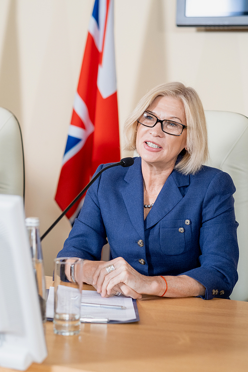 Confident mature blond female delegate making report at political conference while sitting in front of audience