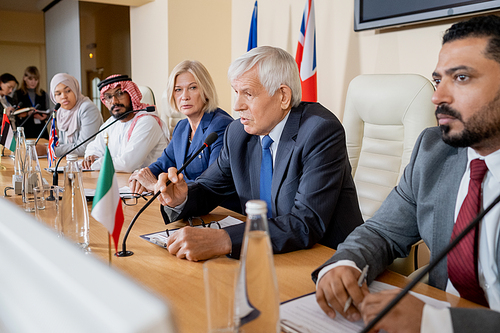 Aged politician in formalwear making report for audience at conference while his foreign colleagues looking at him