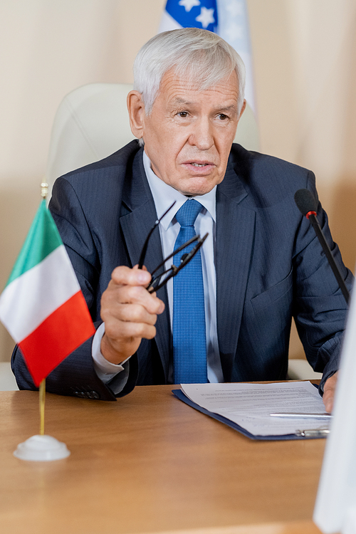 Mature confident politician in suit speaking in microphone while making important report for audience at conference