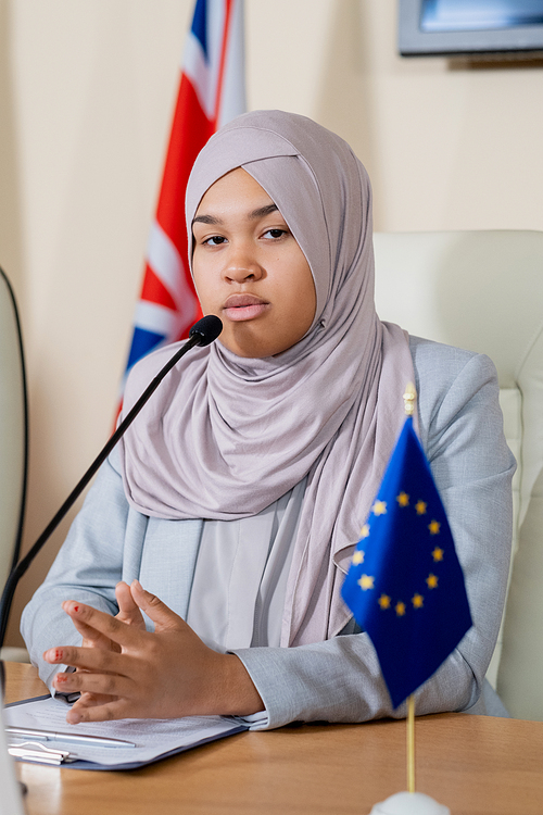 Young successful female politician in hijab sitting in front of camera and looking at you while making report