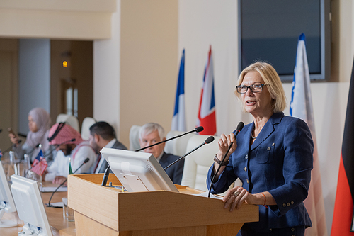 Mature female speaker in elegant suit making report while standing by tribune against row of foreign colleagues