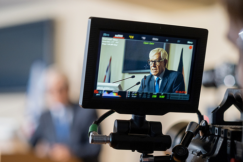 Display of camera with aged male delegate making report at political conference or some other event