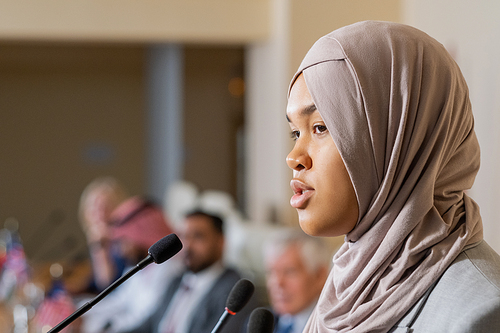 Young pretty female speaker in hijab talking to audience at conference, political forum or summit