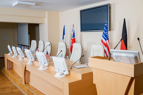 Empty conference hall with row of microphones, computer monitors and flags on long table, tribune and armchairs