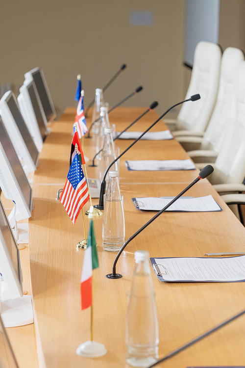 Row of bottle with water, national flags, microphones, computer monitors and papers on long table in conference hall