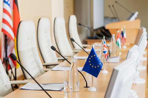 Row of microphones, international flags, bottles of water, glasses, computer monitors and papers on long table in conference hall