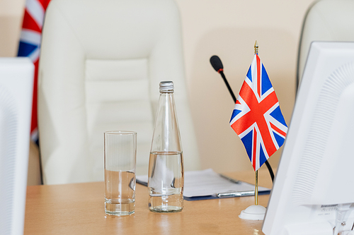 Bottle of water, glass, microphone and British flag on workplace of delegate with white armchair on background