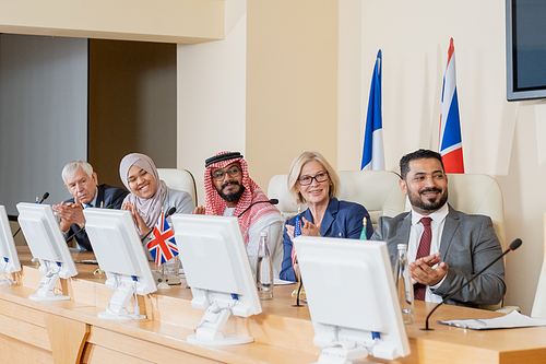 Happy intercultural delegates in formalwear clapping hands to speaker after report while sitting along table in conference hall