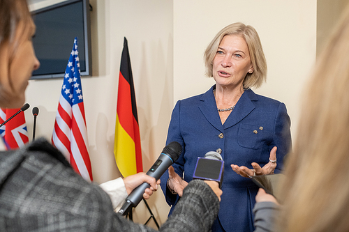 Contemporary female politician in formalwear giving interview to journalists at press conference against national flags