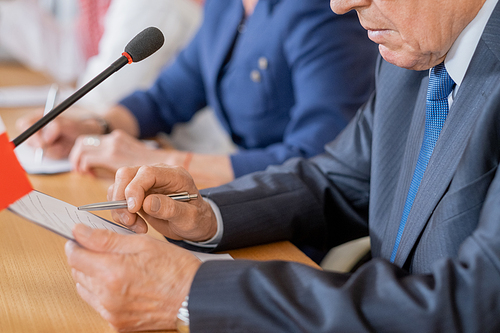 Hands of mature delegate pointing at document in clipboard while making report by table against row of foreign colleagues