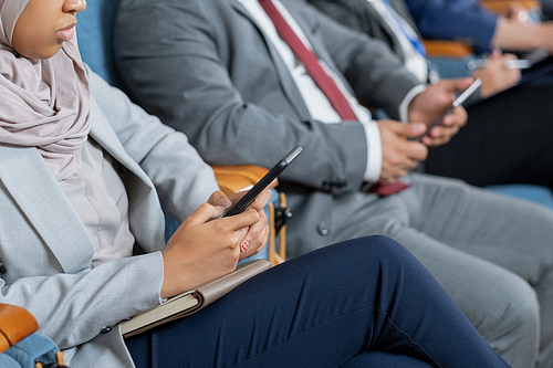 Young Muslim businesswoman scrolling in mobile phone while sitting against row of foreign colleagues