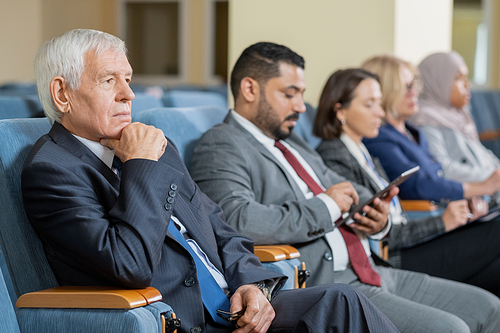 Senior businessman or politician in formalwear listening to speaker at conference while sitting in row with foreign colleagues