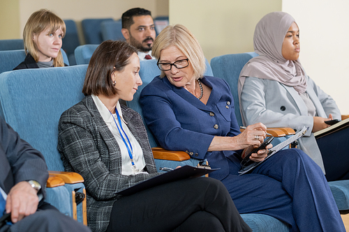 Mature blond female delegate in formalwear talking to young partner at lecture in conference hall