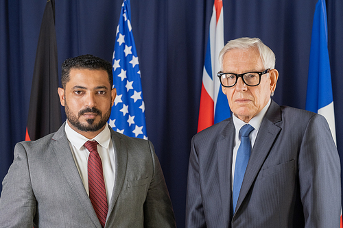 Two elegant male delegates from various countries standing against flags and looking at camera in conference hall