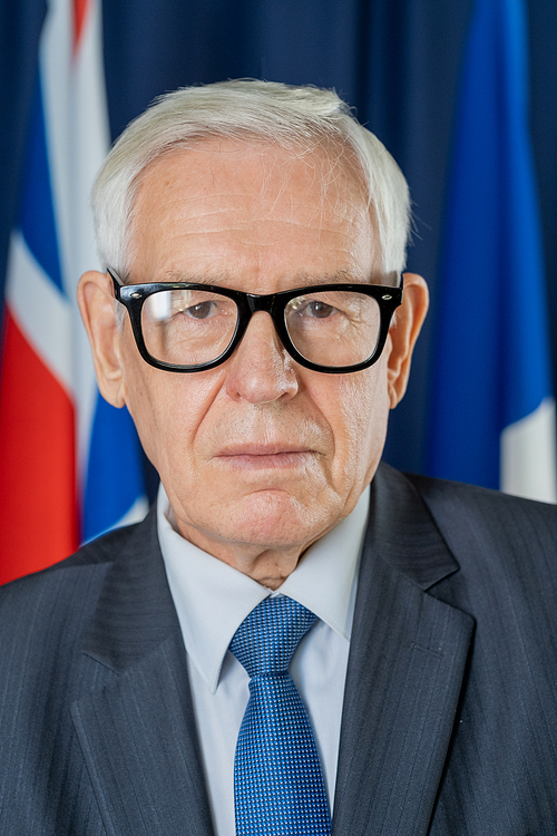 Serious senior politician in eyeglasses and suit standing in front of camera on background of British and Russian flags