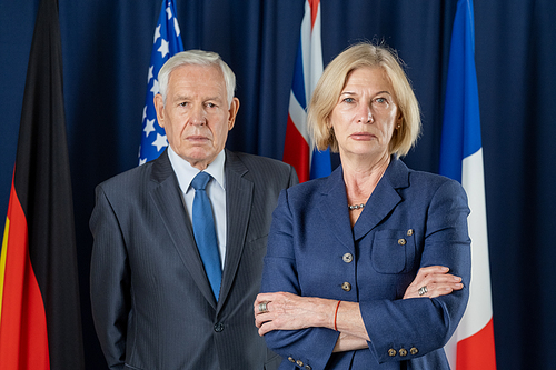 Two mature political leaders in formalwear standing against four flags in conference hall and looking at camera