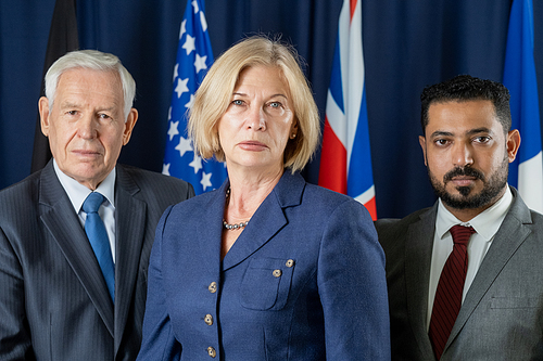 Group of mature male and female politicians in formalwear looking at you while standing against flags