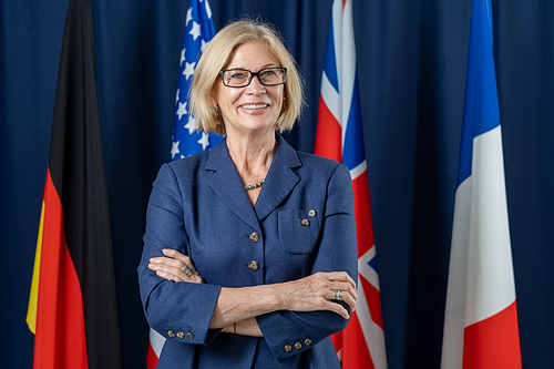 Mature successful political leader crossing arms by chest and looking at camera on background of four flags