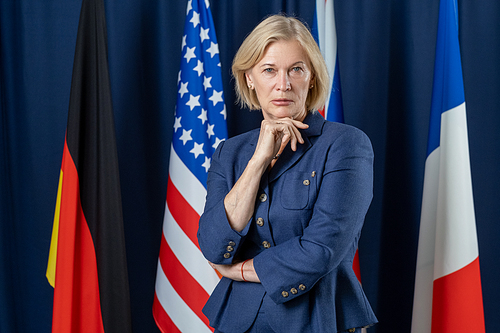 Contemporary mature female delegate in formalwear looking at you while standing against flags of several countries