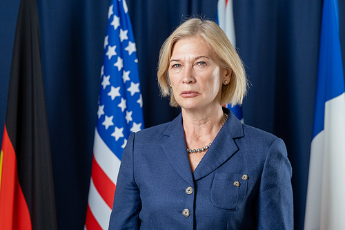 Serious mature female leader in elegant suit standing in front of camera with row of flags on background