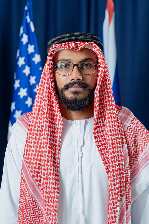 Young serious political leader in national clothes and eyeglasses standing in front of camera against American and Russian flags