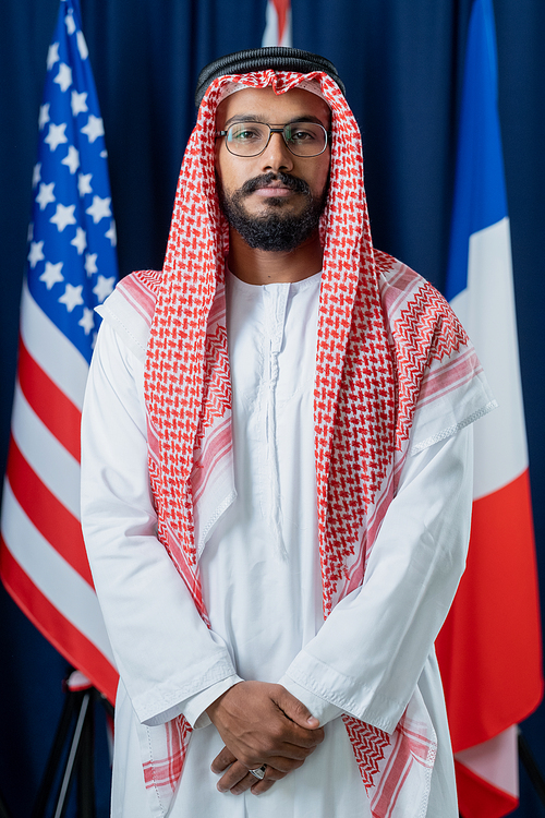 Young Arabian man in turban and national clothes keeping hands put together while standing in front of camera