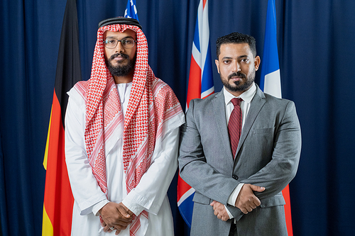 Two intercultural delegates standing against flags of countries included in g7 in conference hall and looking at camera