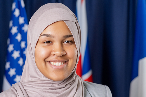 Young contemporary Muslim female delegate in hijab looking at camera with smile while standing against flags